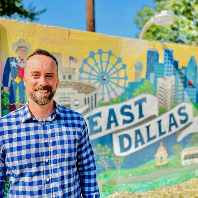 Alex Lowe, smiling in a blue checked shirt in front of a painted East Dallas mural.